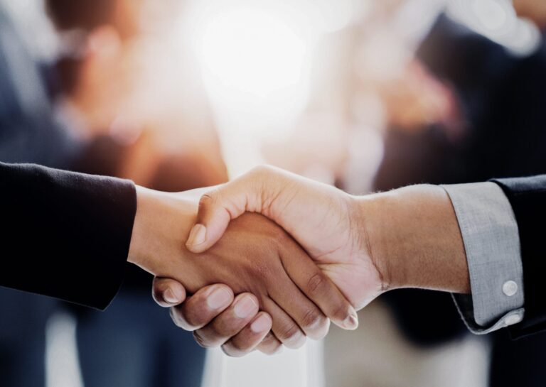 Zwei Personen schütteln sich die Hand, im Hintergrund sind unscharf weitere Menschen zu sehen.